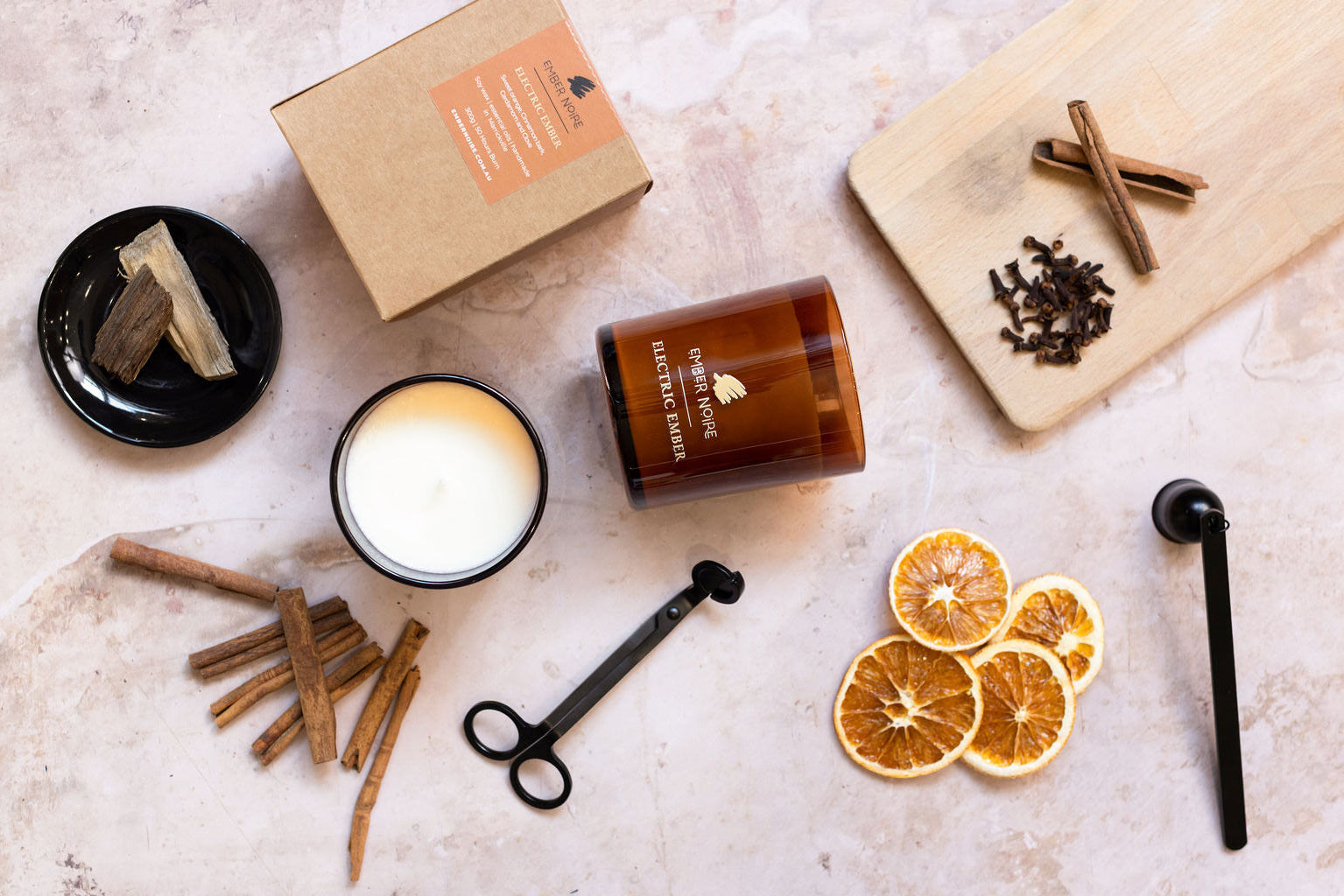 Candle with packaging, cinnamon sticks, and dried oranges on a light surface