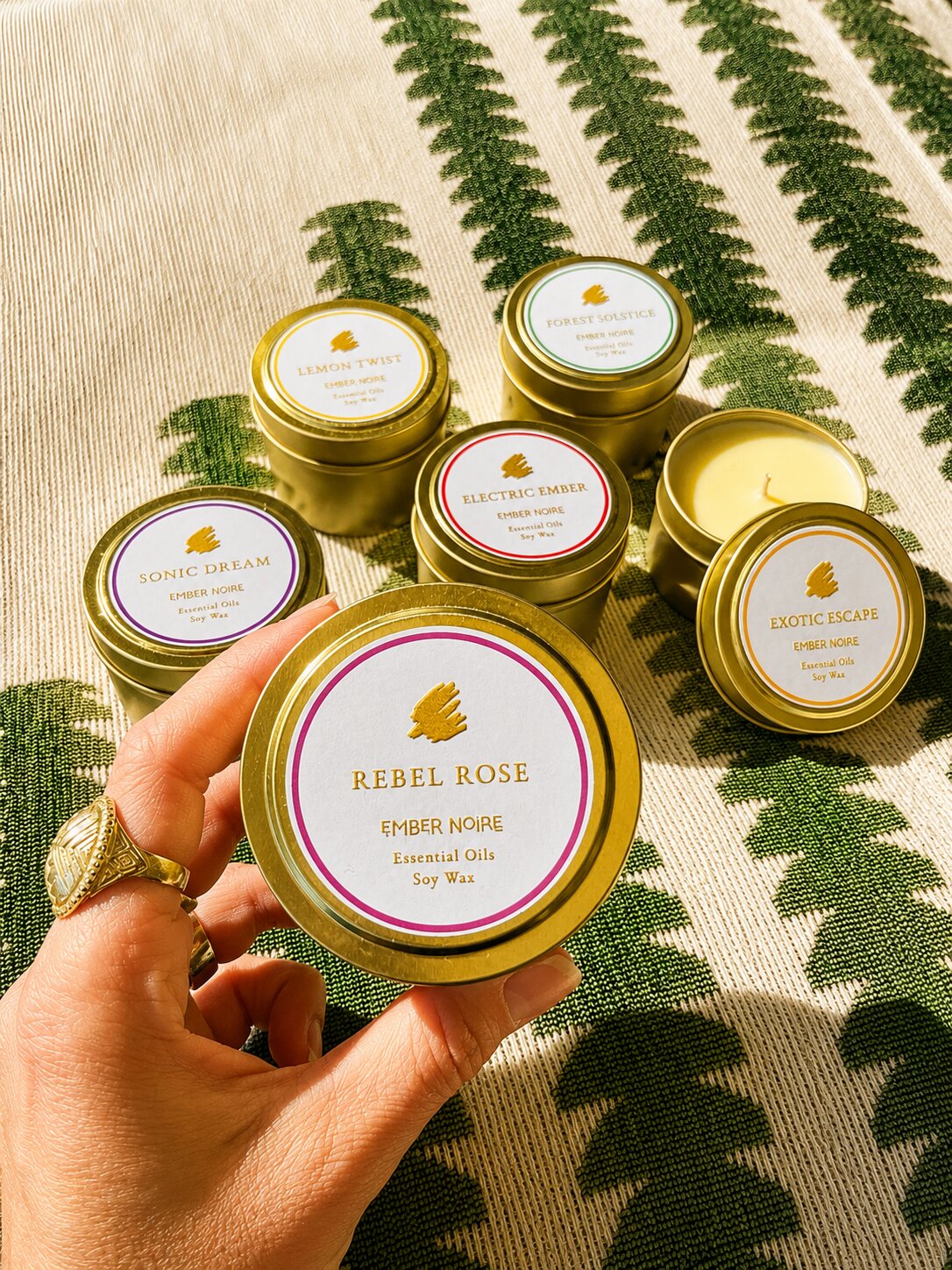 A hand holds a gold tin candle labelled 'Rebel Rose' by Ember Noire, with the other discovery set mini candles arranged on a green and cream patterned textile behind.