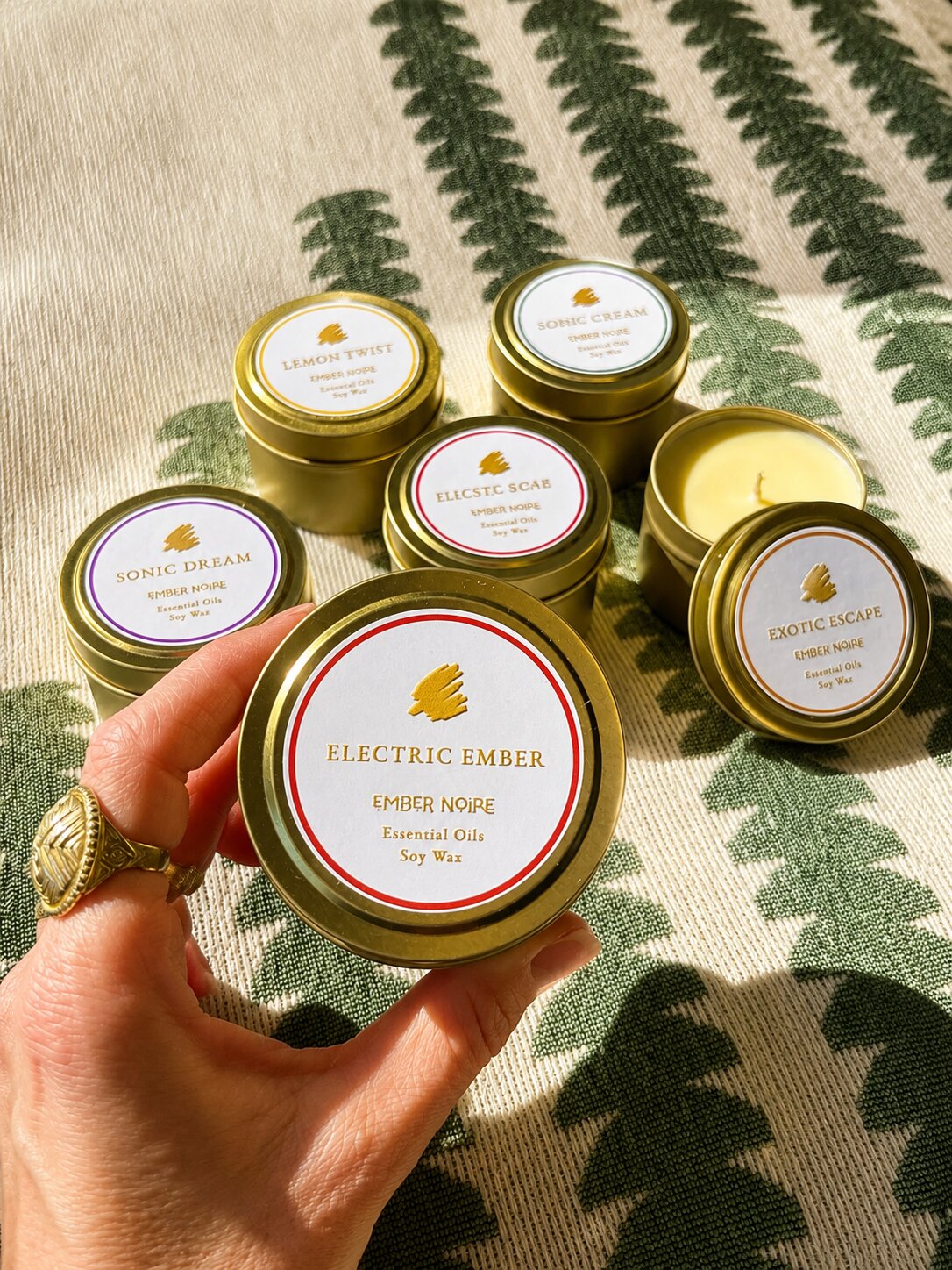 A hand holds a gold tin candle labelled 'Electric Ember' by Ember Noire, with the other discovery set mini candles arranged on a green and cream patterned textile behind.