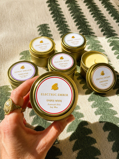 A hand holds a gold tin candle labelled 'Electric Ember' by Ember Noire, with the other discovery set mini candles arranged on a green and cream patterned textile behind.