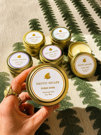 A hand holds a gold tin candle labelled 'Exotic Escape' by Ember Noire, with the other discovery set mini candles arranged on a green and cream patterned textile behind.