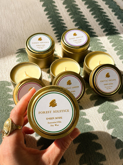 A hand holds a gold tin candle labelled 'Forest Solstice' by Ember Noire, with the other discovery set mini candles arranged on a green and cream patterned textile behind.