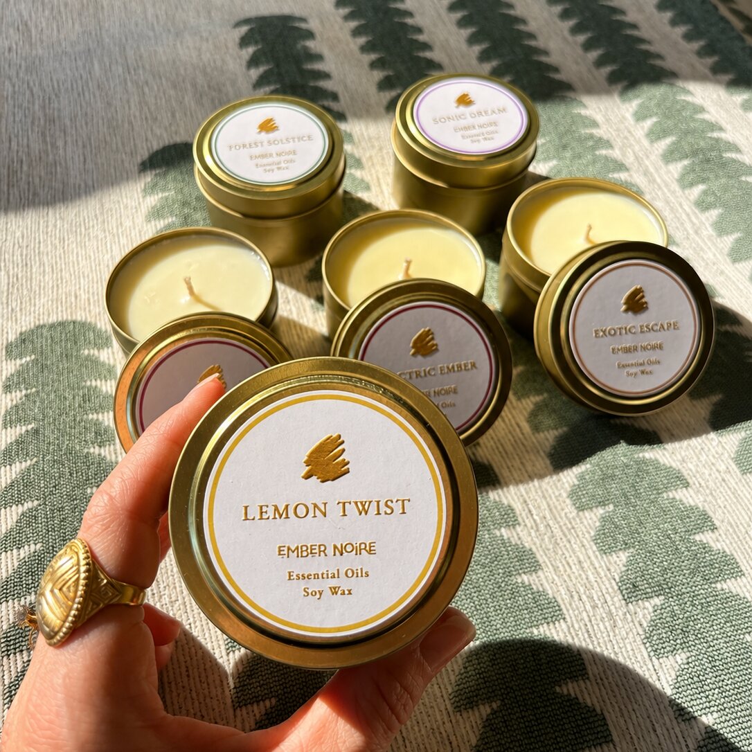 A hand holds a gold tin candle labelled 'Lemon Twist' by Ember Noire, with the other discovery set mini candles arranged on a green and cream patterned textile behind.