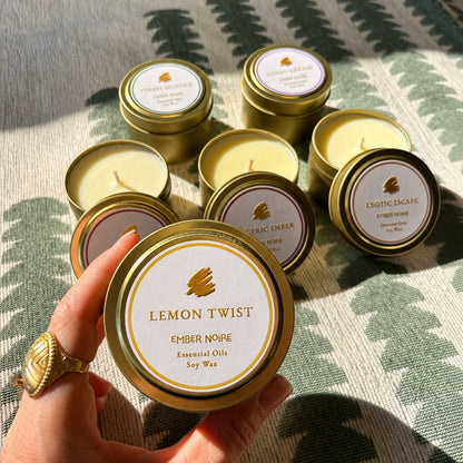 A hand holds a gold tin candle labelled 'Lemon Twist' by Ember Noire, with the other discovery set mini candles arranged on a green and cream patterned textile behind.