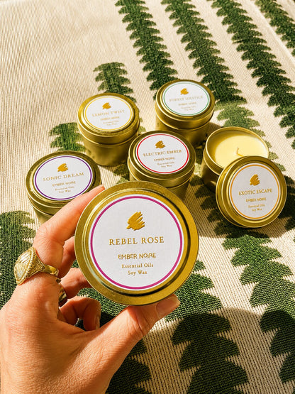 A hand holds a gold tin candle labelled 'Rebel Rose' by Ember Noire, with the other discovery set mini candles arranged on a green and cream patterned textile behind.