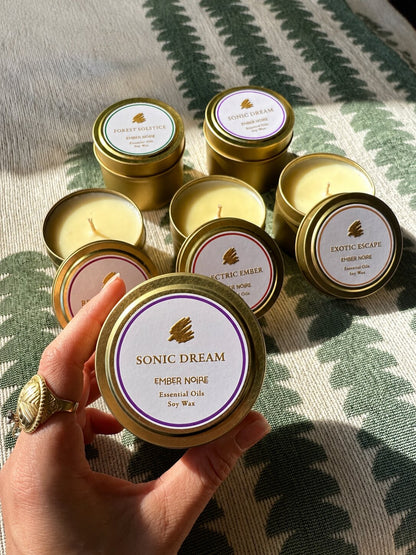 A hand holds a gold tin candle labelled 'Sonic Dream' by Ember Noire, with the other discovery set mini candles arranged on a green and cream patterned textile behind.